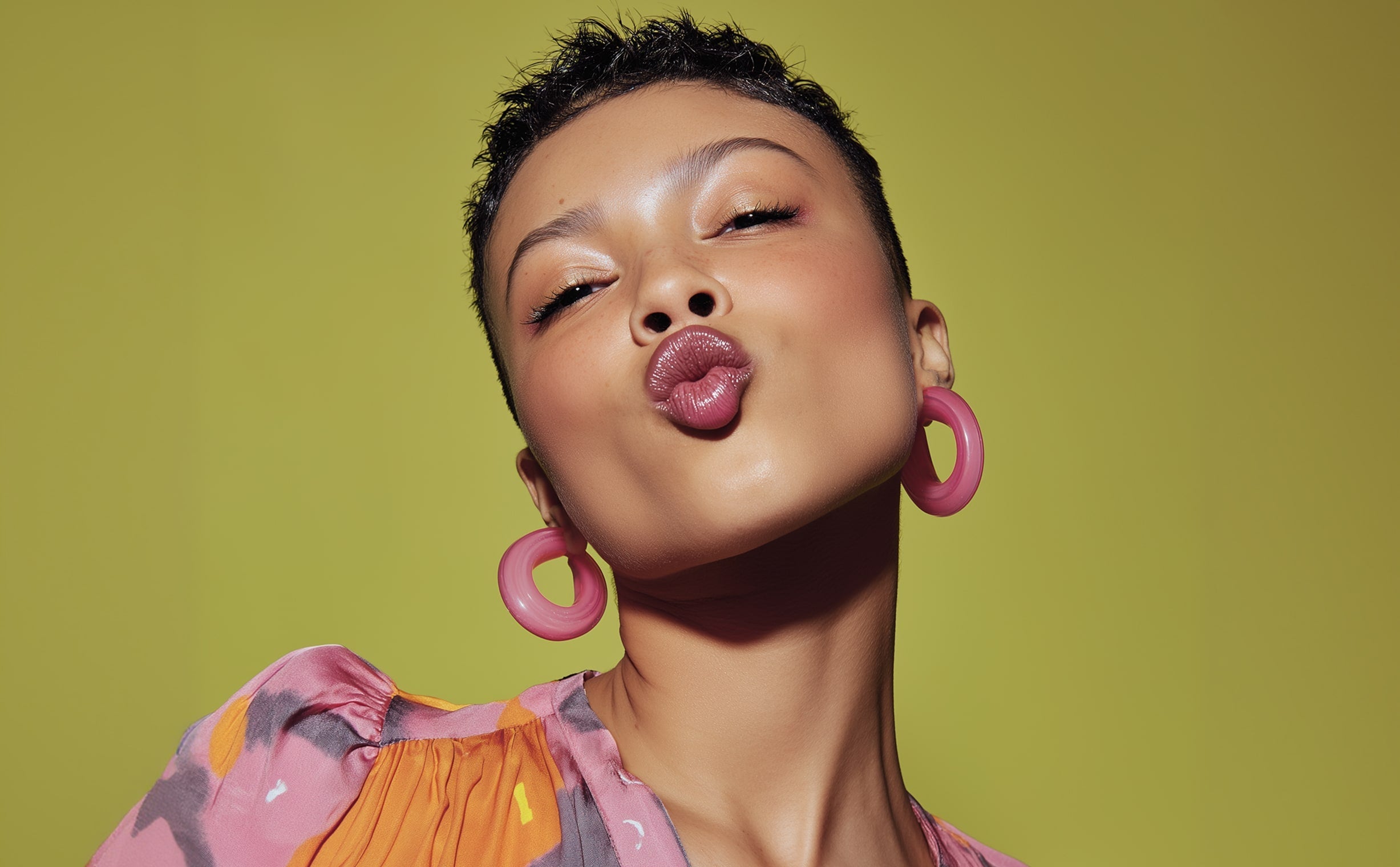 Portrait of woman with short hair wearing pink hoop earrings and colorful outfit against olive background