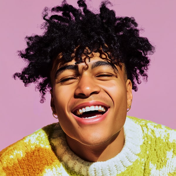 Smiling young man with curly hair wearing colorful knit sweater against pastel pink background