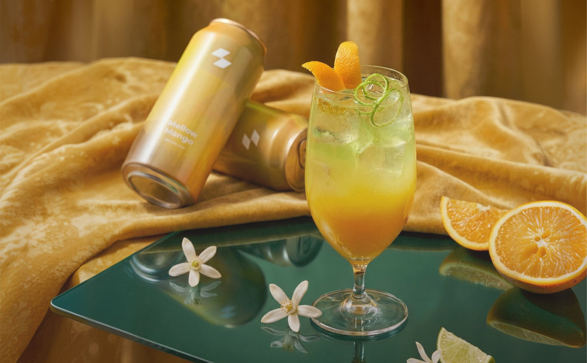 Citrus cocktail with orange garnish beside sparkling wellness drink cans on elegant golden fabric background