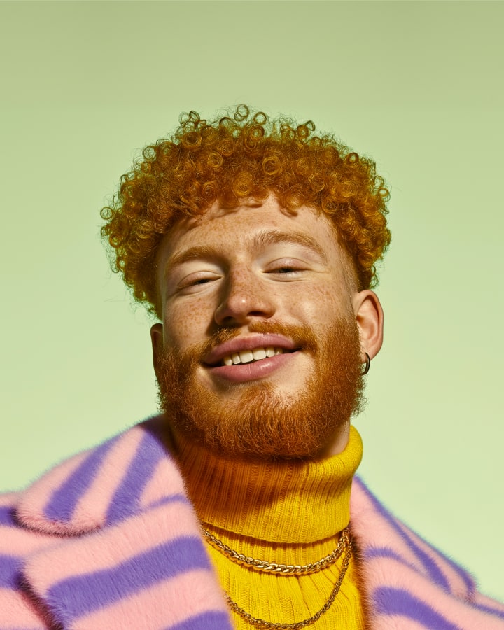 Person with curly red hair and a beard wearing a yellow sweater and striped scarf against a light green background