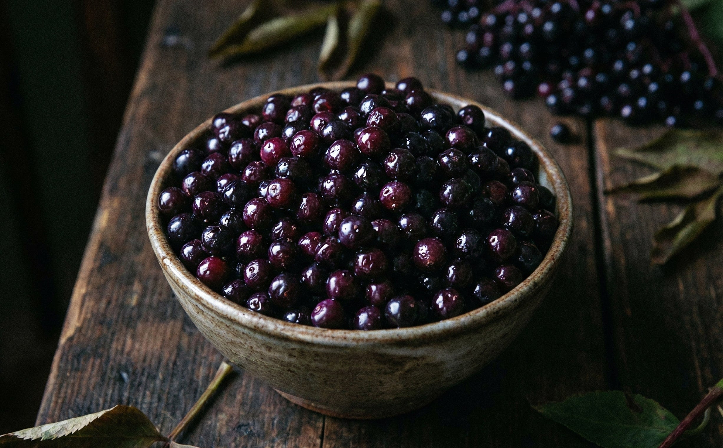Bowl of dark purple berries on a wooden surface with leaves.