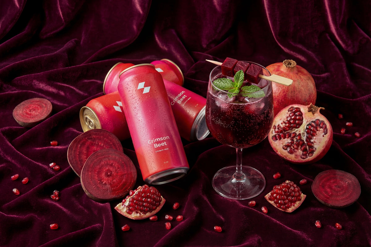 Crimson Beer cans and a glass of red beverage with pomegranate slices on a dark purple fabric background.