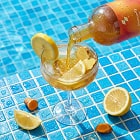 Person pouring a drink into a glass with lemons on a poolside table.
