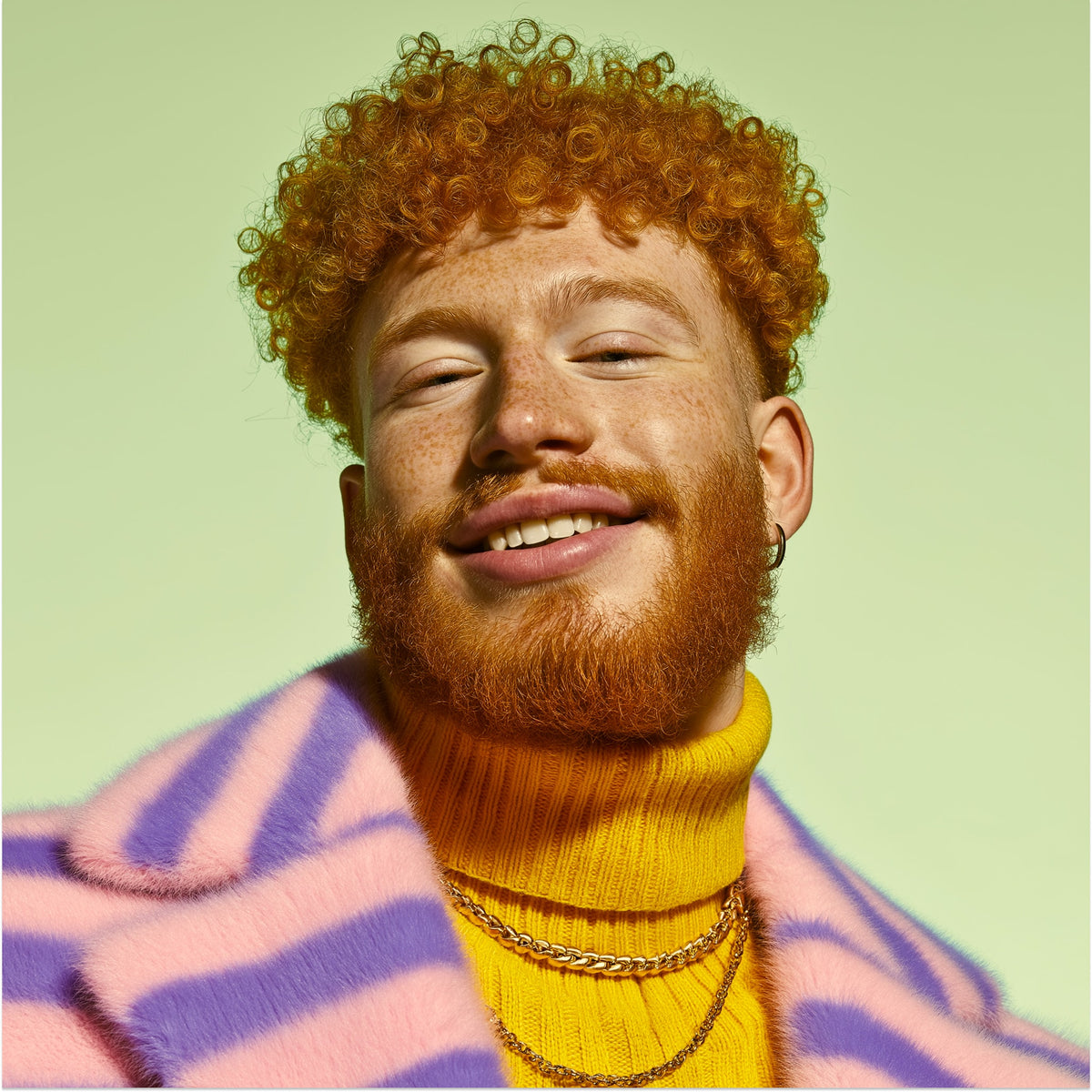 Man with red hair and beard wearing a yellow turtleneck and colorful sweater against a light green background